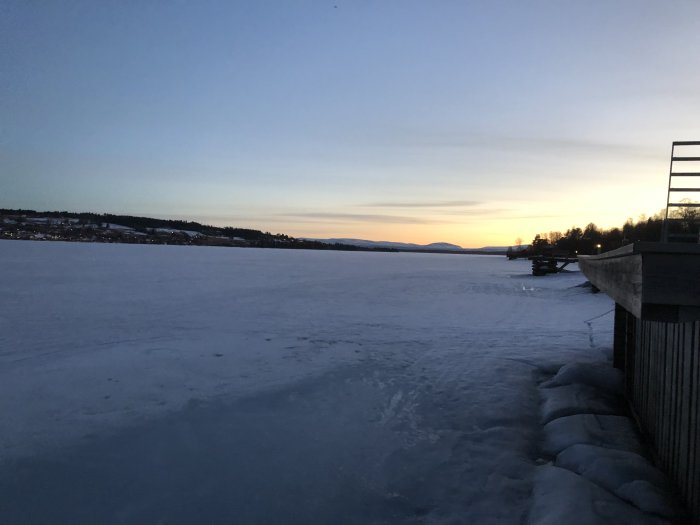 Vinterlandskap vid skymning med snötäckt frusen sjö och byggnad till höger.