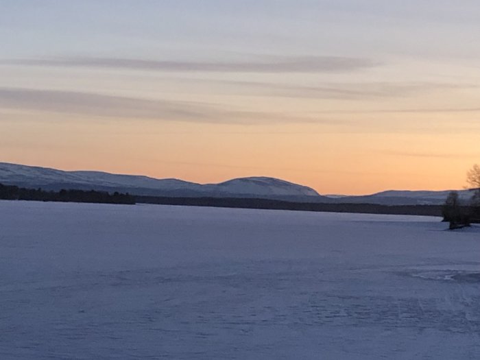 Vinterlandskap vid skymning med snötäckt mark och berg i bakgrunden.