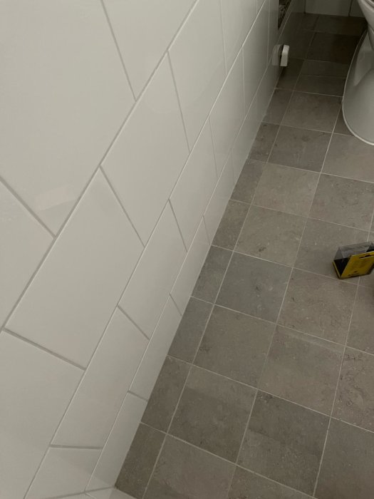 Nyrenoverat badrum med gråa klinkers på golvet och vita kakelplattor på vägg, ej parallella med väggen.