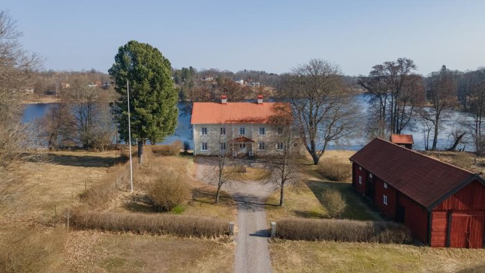 Vy över Skirö Prästgård med ett rött tvåvåningshus, intilliggande röda ladugård, träd och en sjö i bakgrunden.