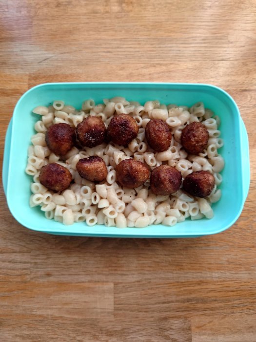 Matlåda med kokt pasta och stekta köttbullar på en träbänk.