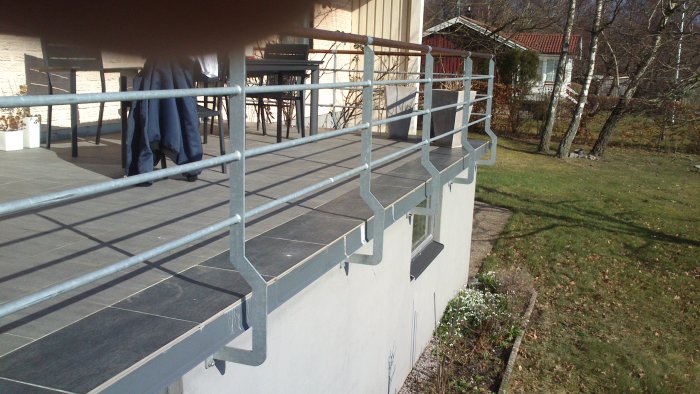 Modern terrass med grå klinkerplattor och metallräcke, med gräsmatta och hus i bakgrunden.