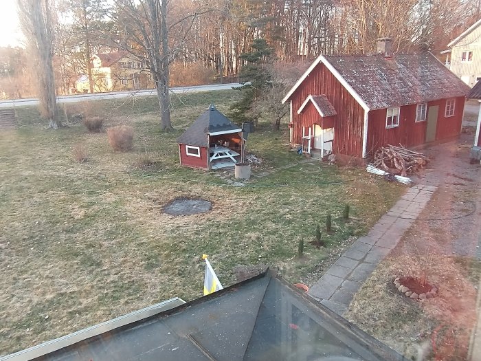 Utsikt från hus över gräsmatta med grillplats och rött skjul, intill en väg och träd.