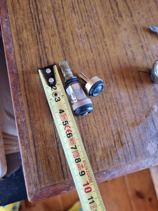 Cylindriskt byggbeslag bredvid ett måttband på ett träbord som visar beslagets storlek i centimeter.