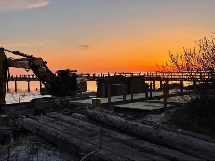 Grävmaskin nära en kaj vid solnedgång, med byggmaterial i förgrunden.