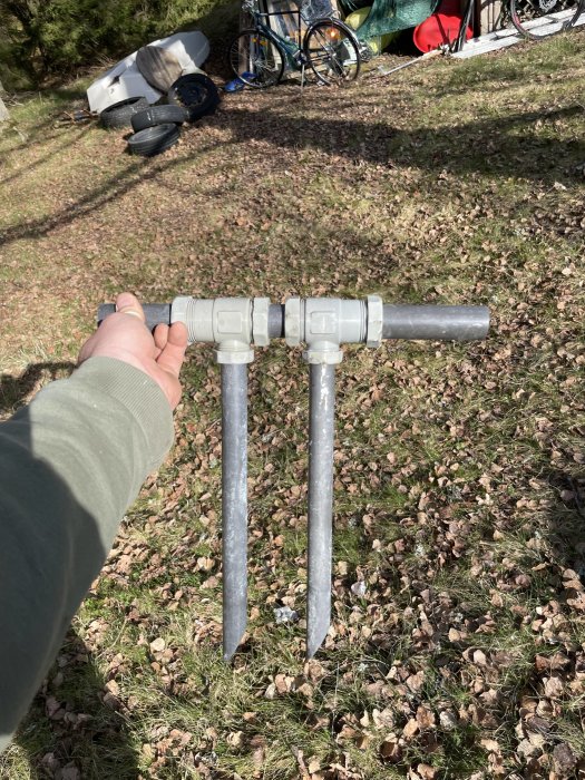 Hand håller en röranordning med två nedåtriktade spetsar mot en gräsmatta med cykel och däck i bakgrunden.