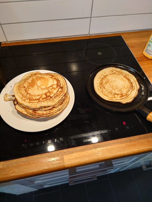 Stapel med nylagade pannkakor på en tallrik bredvid en stekpanna på en spis.