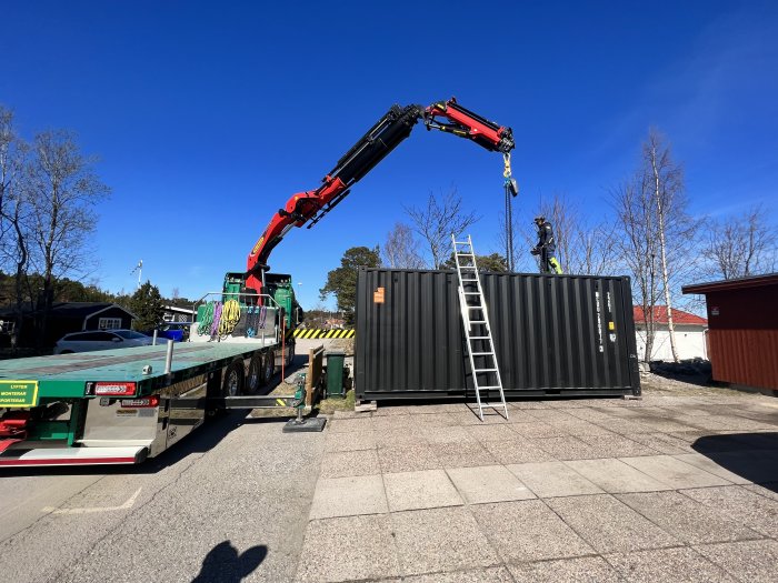 Lastbil med kran lyfter en stor container bredvid en stege och en person.