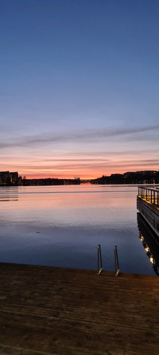 Solnedgång över lugnt vatten sett från träbrygga med stegar och upplyst handräcke.