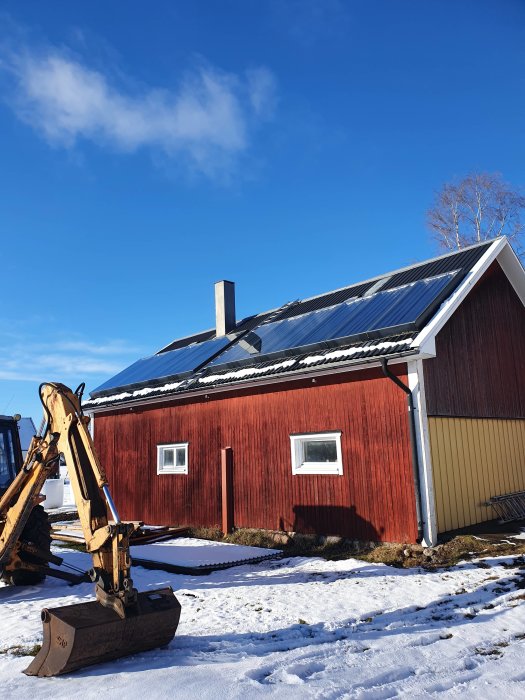 Garage med solpaneler på taket och en grävmaskin i snöklädd omgivning under en klarblå himmel.