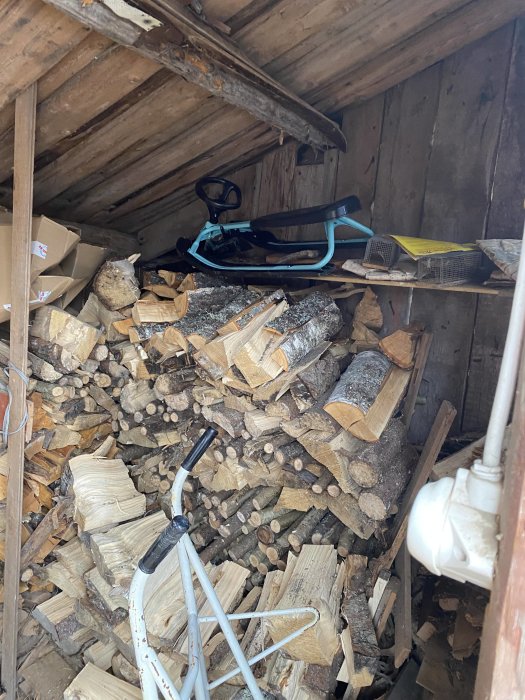 En vedbod full med stablet ved och några utspridda föremål, inklusive en pulka och sopborste.