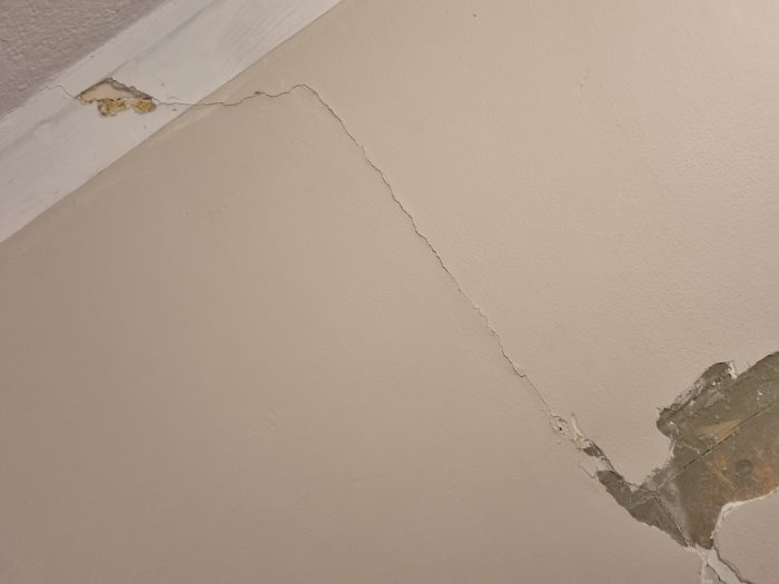 Djup spricka i vitmålad vägg med skadat område nära taklist och delvis borttagen tapet.