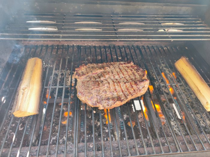 Stor köttbit grillad med tydliga ränder på en kolgrill med glödande kol och rökved.