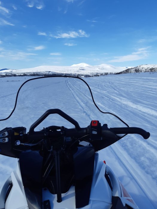 Vy från en snöskoters framdel med spår i snön framåt mot snöklädda berg och klarblå himmel.