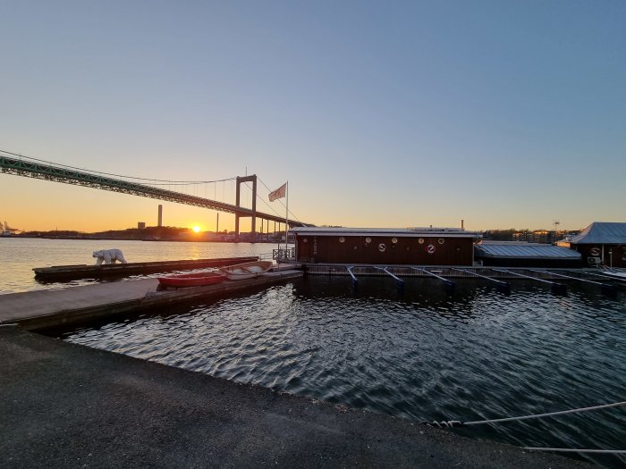 Solnedgång bakom en bro med båthus och småbåtar vid vattnet.