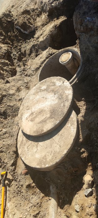 Grävd grop som visar äldre betongbrunnar med lock och rör, halv meter under marken vid dränering.