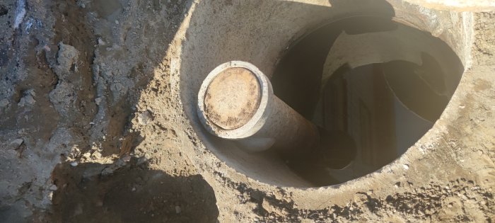 Öppen avloppsbrunn med gammalt tegelrör och lock, halv meter under marken vid dräneringsarbete.