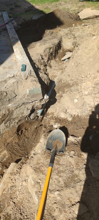 Uppgrävd mark med gammalt tegelrör synligt, spade i förgrunden och schakt för dränering.
