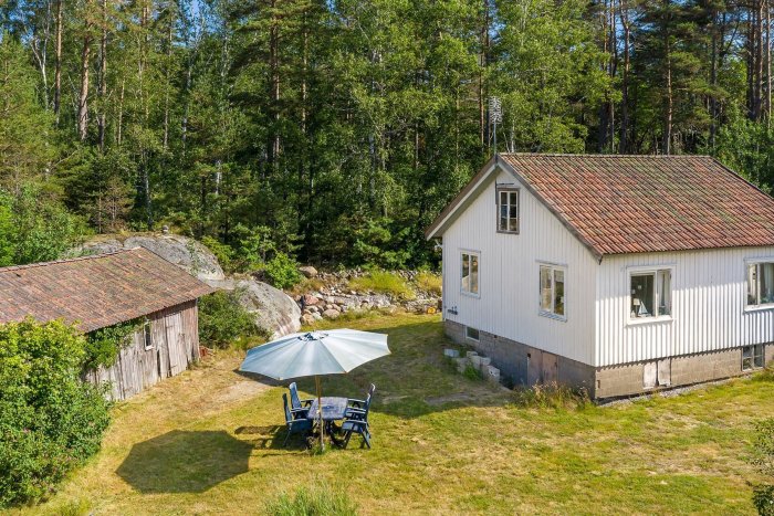 Renoverat vitt torp med oputsad grund, i grönt landskap med trädgårdsmöbler och gammalt skjul.