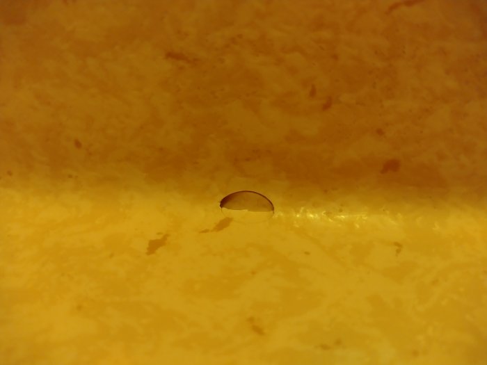 Hål i gult våtrumsmatta med spricka längs den nedre kanten där mattan möter väggen.