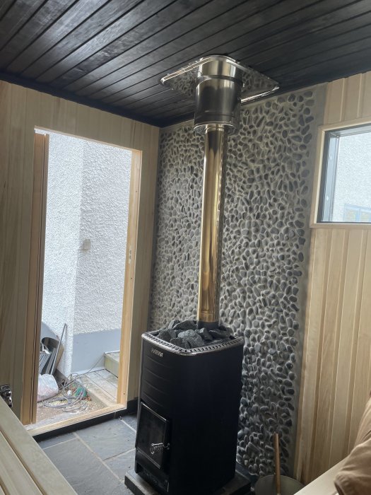 En modern vedeldad bastuugn med skorsten installerad i ett hörn av ett rum med träpanel och stenvägg.