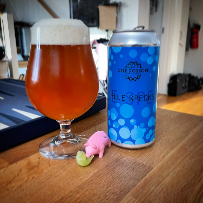 Ölglas fyllt med kopparfärgad dryck bredvid en burk med texten 'Blue Specks' och en liten rosa plastgris.