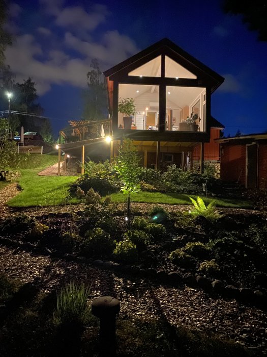 Nattvy av en trädgård med belysning framför ett hus med en inglasad altan och stora fönster svåra att nå för putsning.