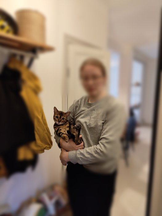 Kvinna med suddigt ansikte håller en bengalkatt i en hall, framför en dörr.