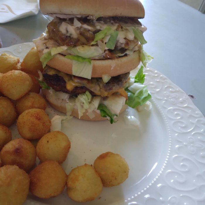 Stor hamburgare staplad med flera köttlag, sås och sallad på tallrik med potatiskroketter.