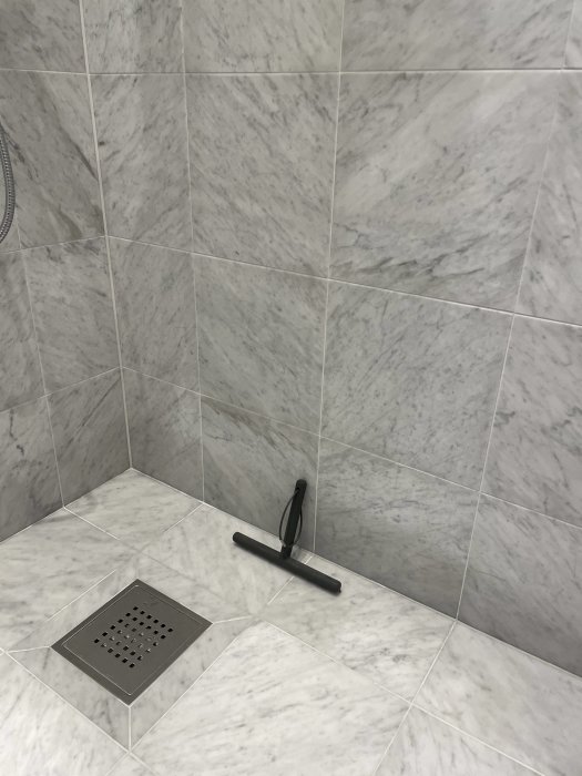 Marmorklätt badrum med mörka fläckar längs väggen och duschskrapa på golvet.