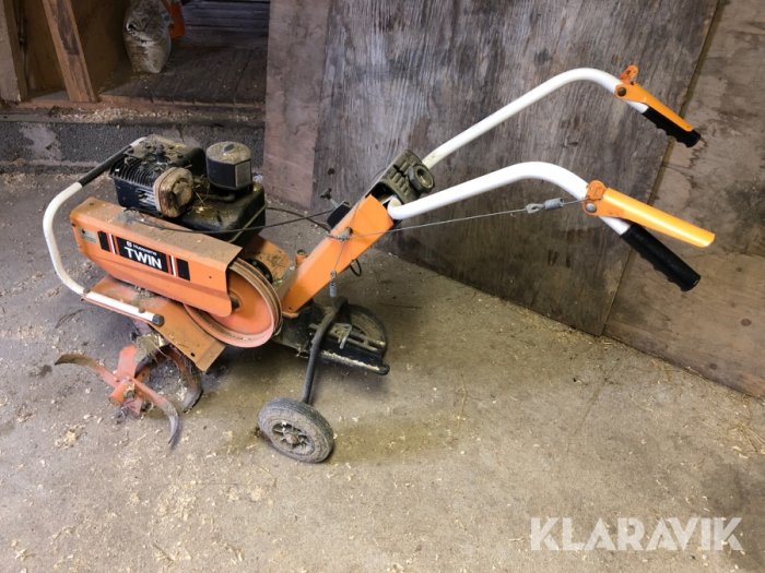 Gammal Husqvarna jordfräs med vit och orange färgschema, saknar en rem och har skadad vajer, parkerad i ett skjul.