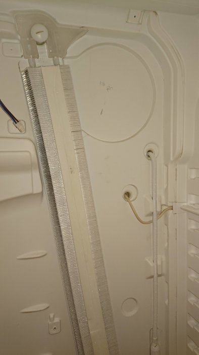Inre ryggpanel av kylskåp med isbildning och synliga kylflänsar och rör.