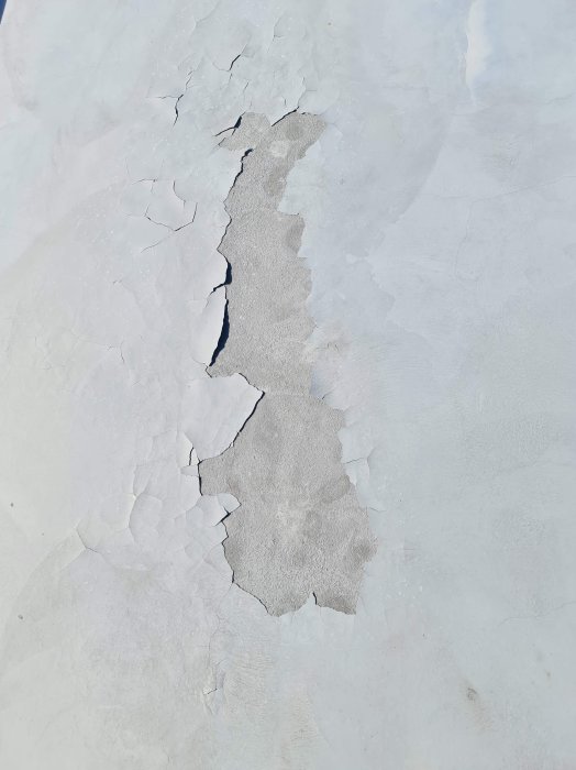 Skadat ytskikt på betongplatta med flagnande och sprucken vit färg som exponerar grå betong undertill.