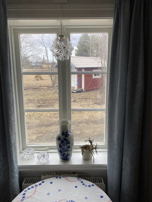 Utsikt från ett oputsat tvåglasfönster i ett äldre hus, med kristallprydnader på fönsterbrädet och en lantlig gård i bakgrunden.