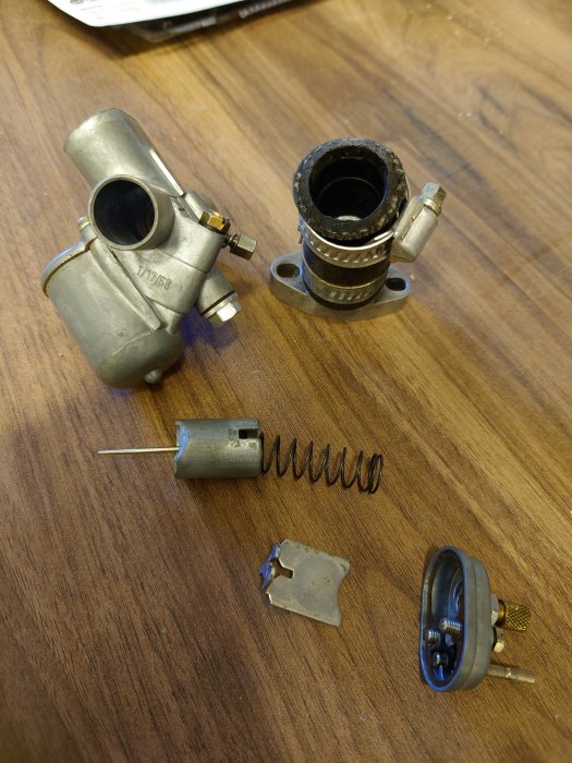 Förgasardelar upplagda på ett träbord, inklusive kropp, munstycke, membran och fjädrar.