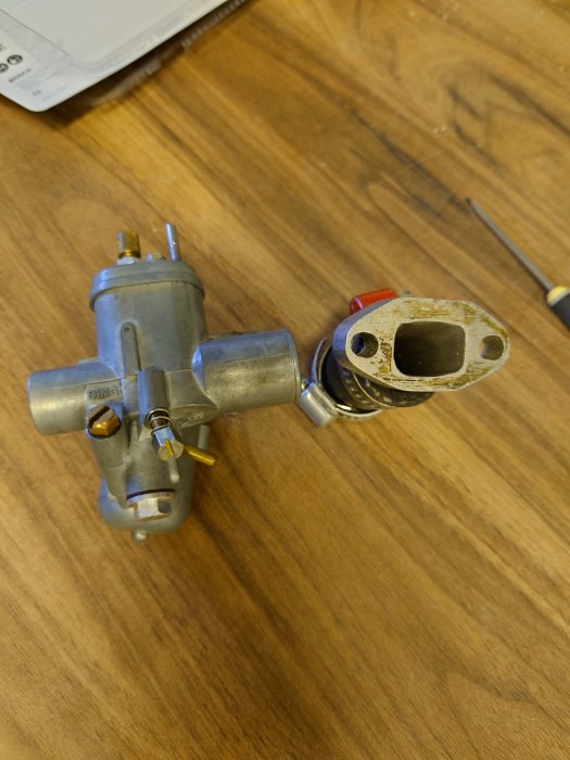 Demonterad förgasare på träunderlag med justeringsskruvar synliga.