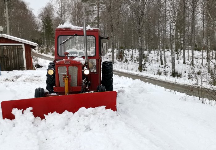 Röd Volvo traktor med plogblad framför snötäckt väg och skog i bakgrunden.