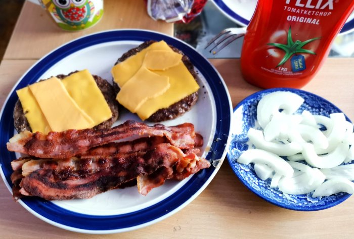 Hamburgare med cheddarost och bacon på en tallrik bredvid lökringar och ketchupflaska.