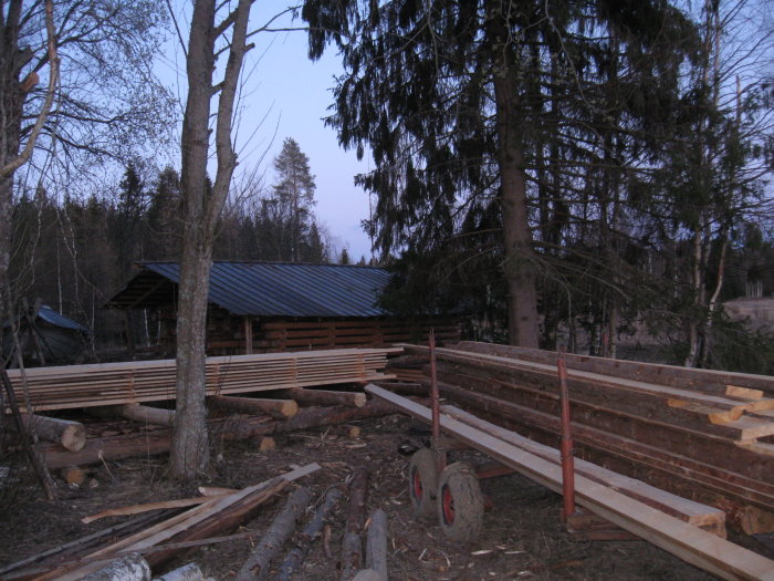 Uppstaplade sågade trästockar utomhus med sågverksutrustning och en skogsbakgrund.