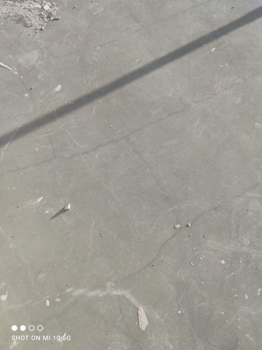 Betonggolv med sprickor och oregelbundna mönster, inklusive en lång skugga som sträcker sig diagonalt över ytan.