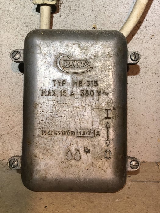 Gammal metallisk elektrisk dosa märkt "Cewe MB 315" på en smutsig vägg med synlig kabel.