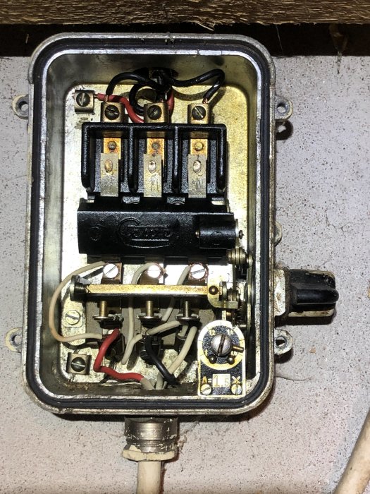 Äldre eluttag med öppen kapsling som visar säkringar, kablar och anslutningar, monterat på en betongvägg.