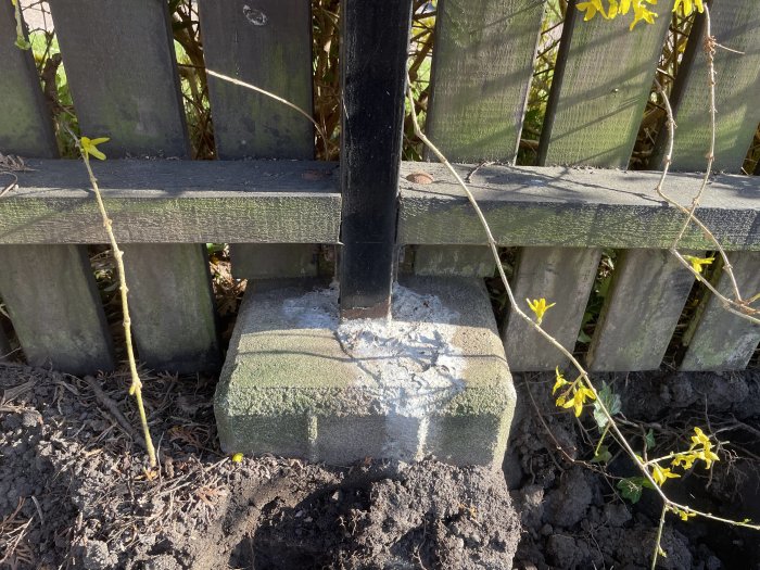 Stålrörsstolpe ingjuten i en stor betongplint vid ett plank, med tecken på rost och slitage.