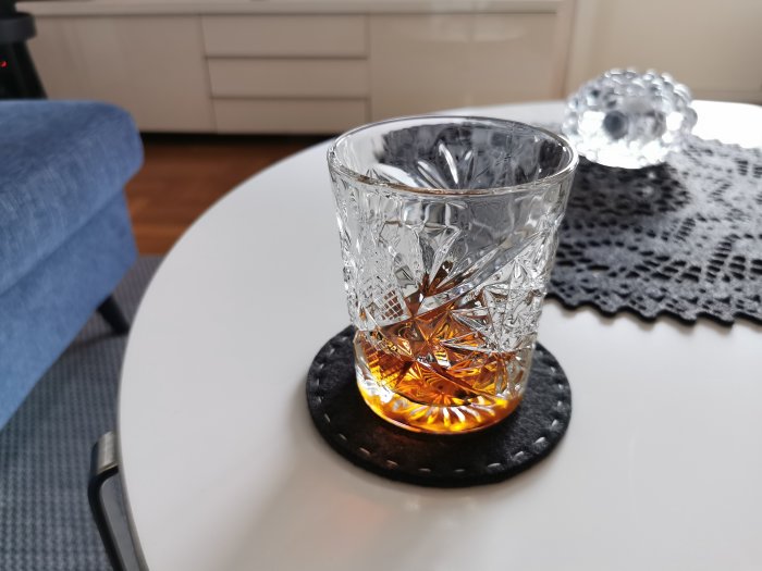 Ett glas med mörk whisky på ett vitt bord, bredvid en blå soffa och en duk.