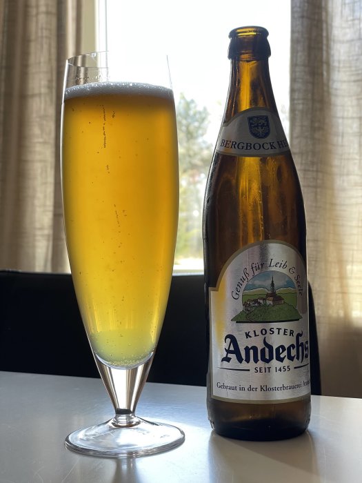 Ett glas med gyllene öl och en tom ölflaska med etiketten "Kloster Andechs" mot fönsterbakgrund.