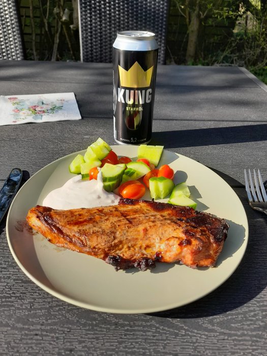 Halv flintastek på tallrik med gräddsås och sallad, brevid en ölburk, utomhus på bord.