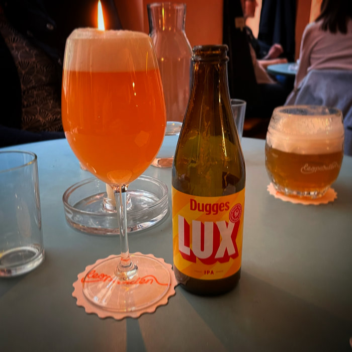 Ett glas öl och en flaska Dugges LUX IPA på bord med levande ljus och suddiga personer i bakgrunden.