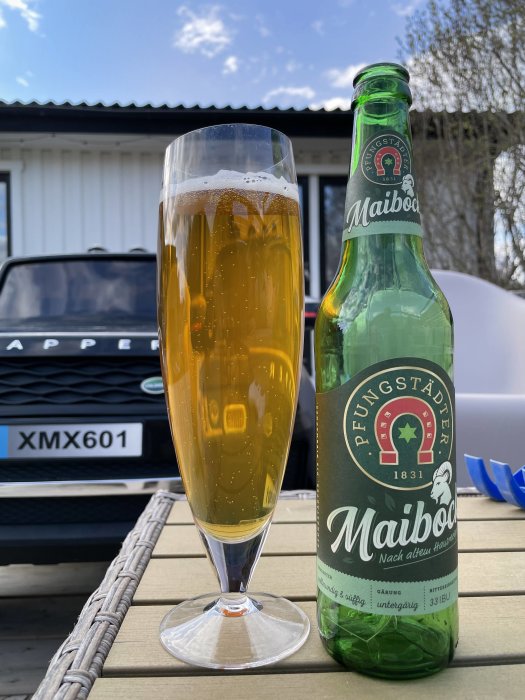 Ett glas med öl och en halvtom Maibock-flaska på ett utomhusbord, med bil och hus i bakgrunden.