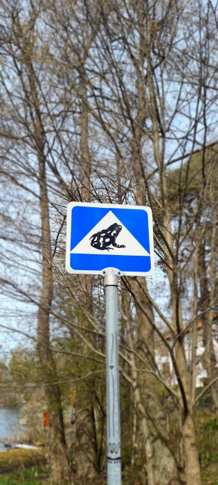 Varningsskylt för grodpassage mot bakgrund av lövfällande träd och himmel.
