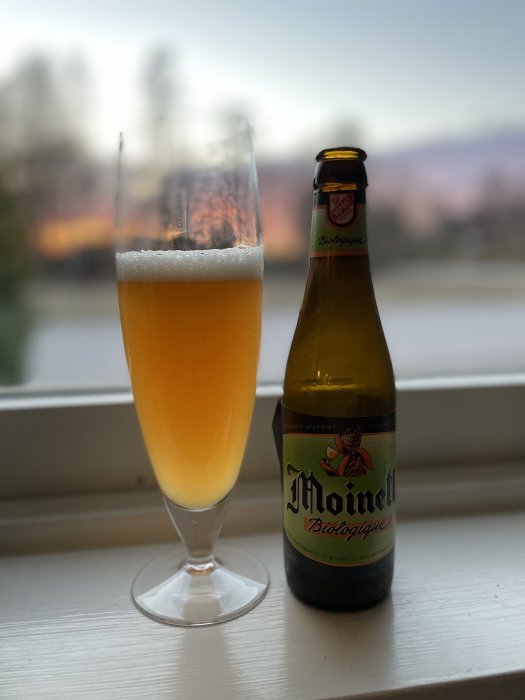 Ett glas fyllt med ljus öl framför en öppen flaska Moinette Biologique på en fönsterbräda med suddig bakgrund vid skymning.
